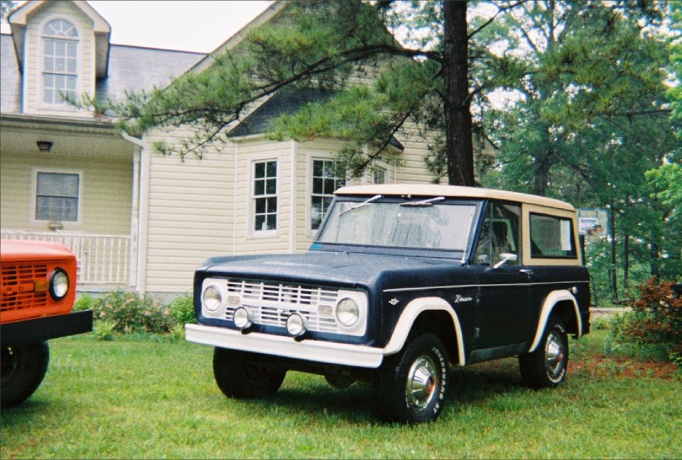 Dark blue Ford Bronco with tan top parked on grass next to an orange vehicle