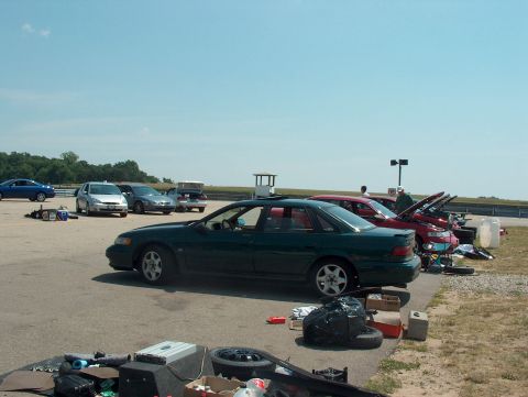 1998 Ford Taurus - A dark green 1998 Ford Taurus is parked on a track alongside other cars and equipment under a clear blue sky.