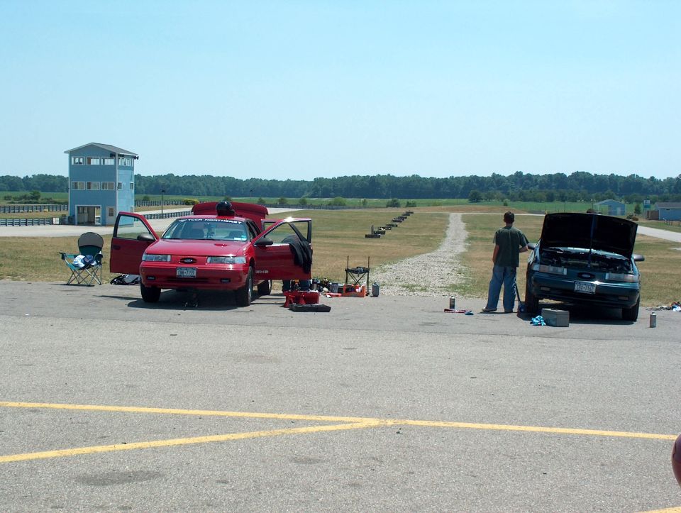 red and dark sedans with hoods and doors open at racetrack pit area