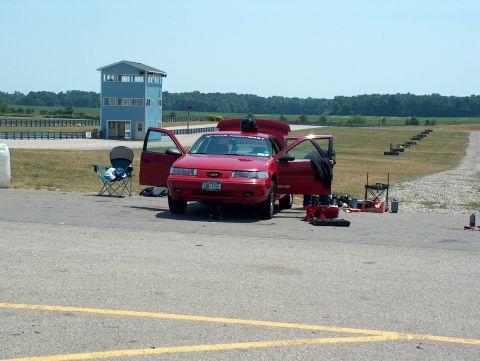 1998 Ford Taurus - A red 1998 Ford Taurus is parked at a racetrack with its doors and hood open, ready for an event.