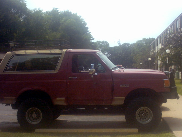 Red lifted Ford Bronco in profile with roof rack, tan hardtop and white-spoke wheels