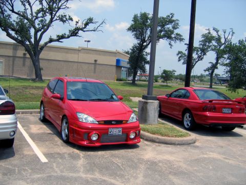 2000 Ford Crown Victoria - A red Ford Focus hatchback is parked next to a red Ford Mustang in a parking lot.