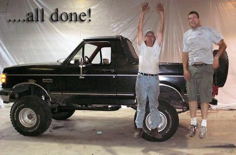 1988 Ford Bronco - Two men stand celebrating beside a freshly painted black lifted full-size Ford Bronco with the top removed, an interior roll bar and white…