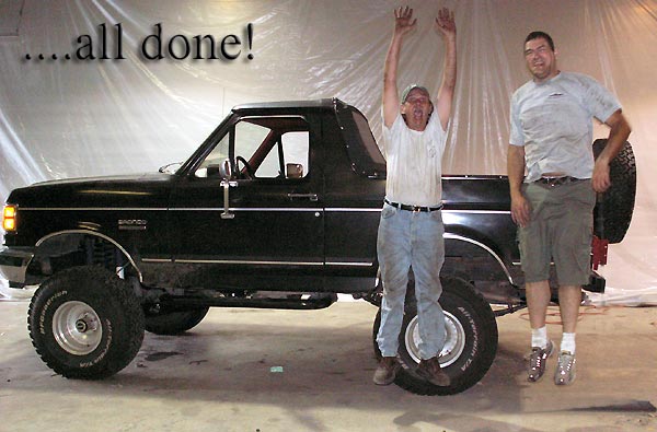Two men beside finished black lifted Ford Bronco with roll bar in plastic-draped paint booth