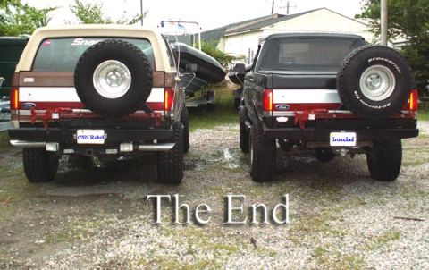 1988 Ford Bronco - Two full-size Ford Broncos photographed from directly behind on a gravel lot, each with a heavy rear bumper and a swing-out spare on a whit…