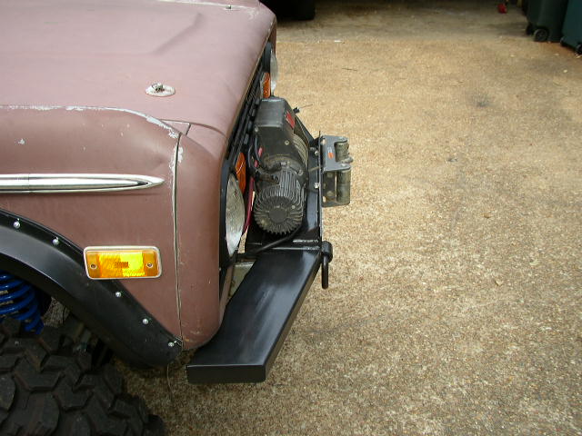 Front corner of a brown Ford Bronco with a winch mounted on the bumper. Shows the headlight, turn signal, and fender flare