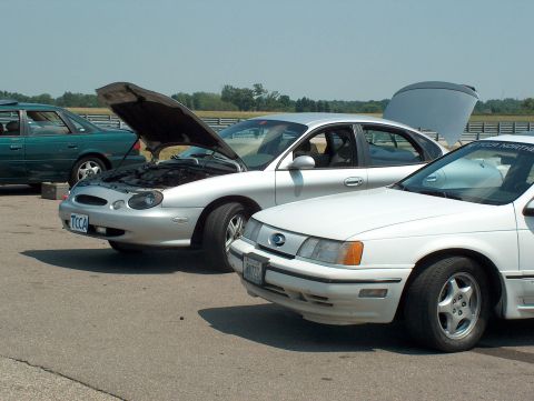 1998 Ford Taurus - Multiple sedans are parked on asphalt, with two having their hoods or trunks open.