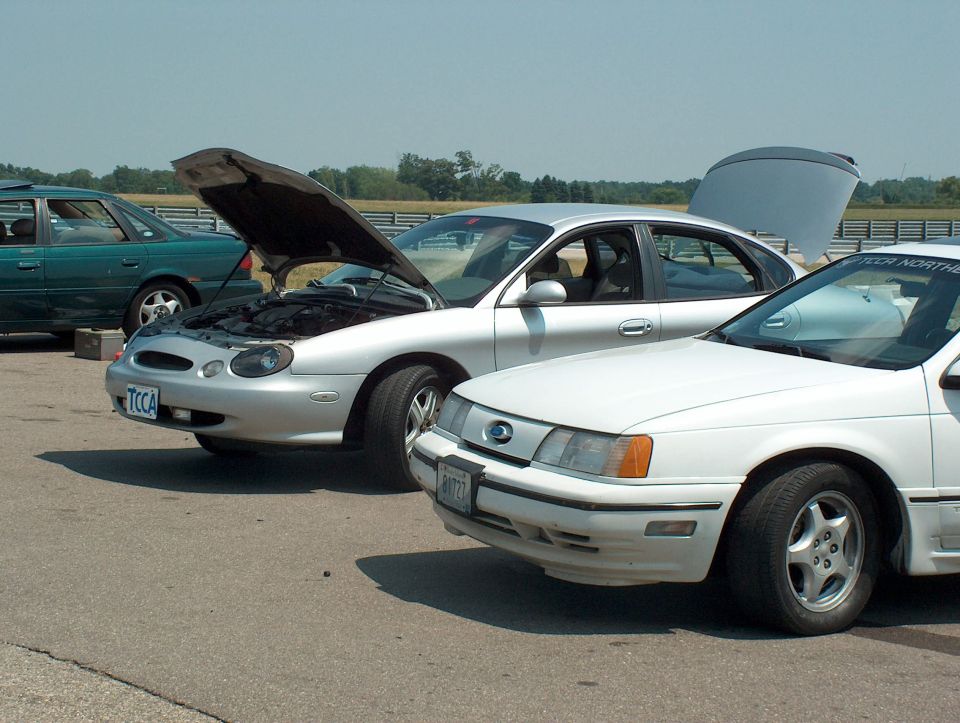 Three sedans parked on asphalt, one silver with hood open, one white with trunk open, and a green one in the background