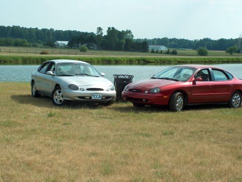 1998 Ford Taurus - Two sedans, a silver 1998 Ford Taurus and a red one, are parked on a grassy area next to a body of water.