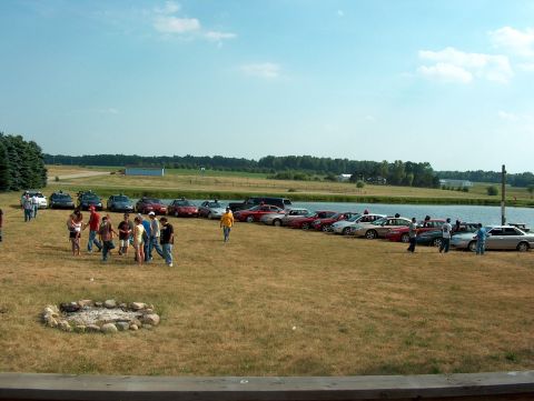 1998 Ford Taurus - A gathering of vehicles and people takes place outdoors near a body of water under a clear sky.