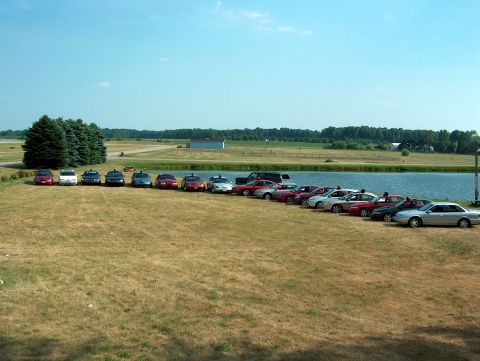 1998 Ford Taurus - A collection of sedans are lined up on a grassy field next to a body of water under a clear sky.