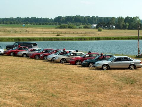 1998 Ford Taurus - Several sedans and an SUV are parked in a row on a grassy field next to a lake.