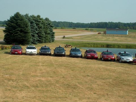 1998 Ford Taurus - A lineup of eight sedans is visible in a grassy field with trees and a pond in the background.