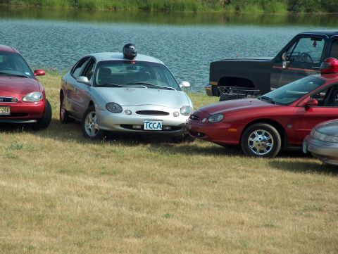 1998 Ford Taurus - Several cars, including a 1998 Ford Taurus with a helmet on its roof, are parked on grass by a lake.