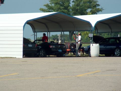 1998 Ford Taurus - Two cars are parked under a white canopy structure at what appears to be a race track or car show.