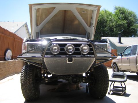 1990 Ford Bronco - lifted truck with its hood up shows a custom tubular front bumper with three round driving lights and large off-road tires.