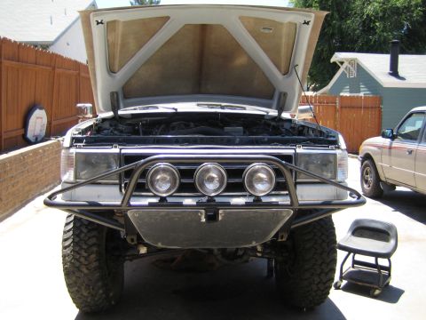 1990 Ford Bronco - lifted pickup with its hood raised sits on a patio, fitted with a tubular front bumper carrying three round driving lights.