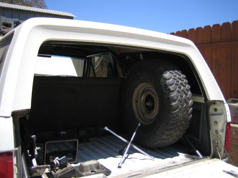 1990 Ford Bronco - open rear cargo area of a white SUV holds a mounted off-road spare tire propped on rods, with tools below in a backyard.