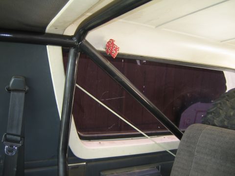 1990 Ford Bronco - vehicle's interior shows a black roll bar at the B-pillar with a red fuzzy-dice ornament, beside a tinted rear quarter window.