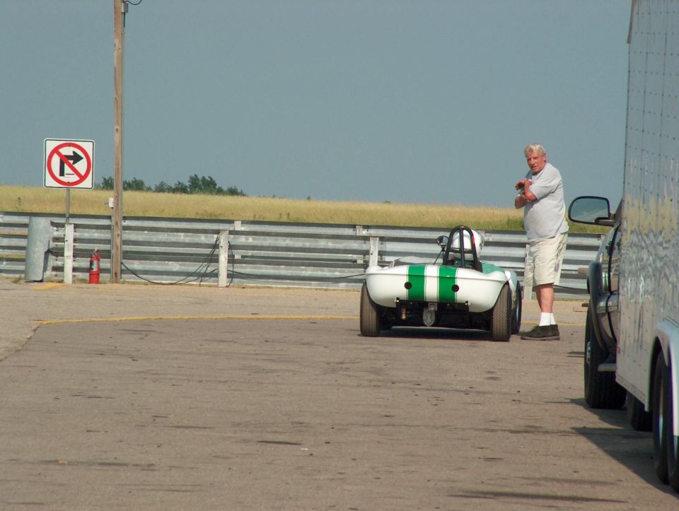 small white race car with green stripes, rear view, parked next to a trailer