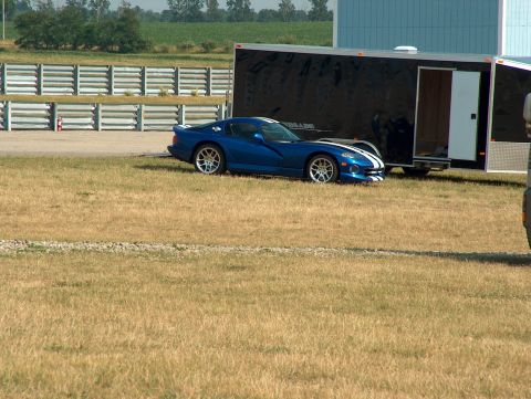 1998 Ford Taurus - A blue sports car with white racing stripes is parked on a grassy field next to a large black trailer.