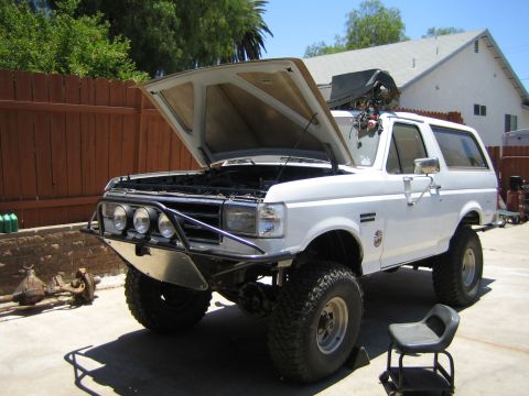 1990 Ford Bronco - white lifted Ford Bronco sits in a backyard with its hood propped open, a tubular front bumper with off-road lights, and large mud tires.