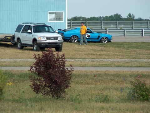 1998 Ford Taurus - A white SUV is parked beside a blue muscle car on a race track, with a person standing near the muscle car.