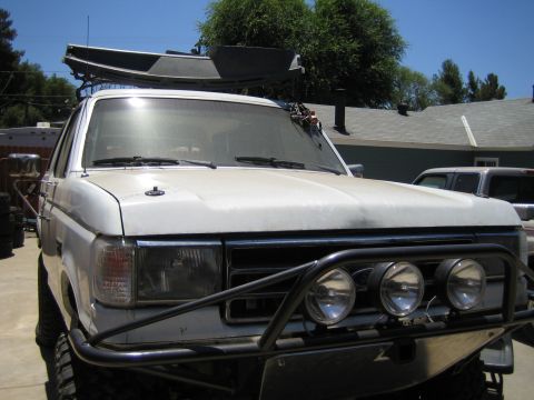 1990 Ford Bronco - front of a white SUV carries a small boat on a roof rack and has a tubular front bumper fitted with three round driving lights.