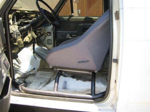 1990 Ford Bronco - bare vehicle interior shows the steering column and dash with a gray fabric racing bucket seat mounted on a tubular floor frame.