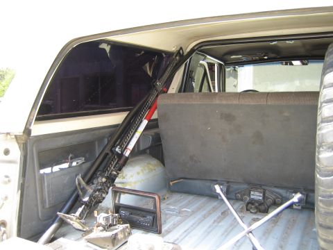 1990 Ford Bronco - rear cargo area of an SUV holds a hi-lift jack, a yellow fuel tank, an X-shaped lug wrench, and a mounted spare tire.