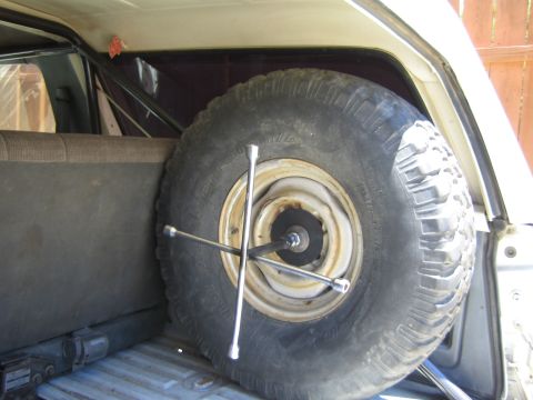 1990 Ford Bronco - mud-terrain spare tire on a white steel wheel is mounted upright in an SUV cargo area with a four-way lug wrench crossing it.