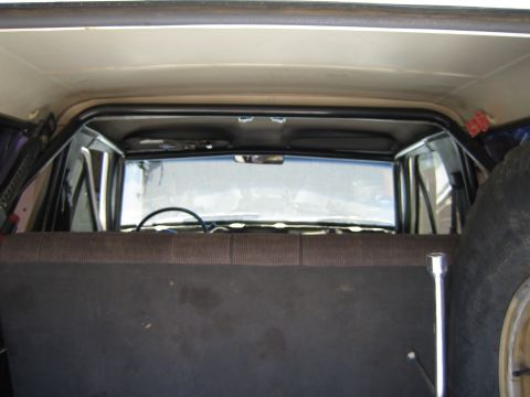 1990 Ford Bronco - Looking forward from the rear of an SUV across a bench seat top toward the rear glass, headliner, and a black roll bar.