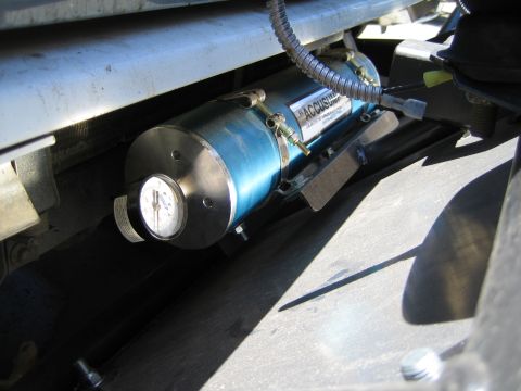 1990 Ford Bronco - blue anodized cylindrical Accusump oil accumulator with a pressure gauge and braided line is mounted to a bracket under a vehicle.