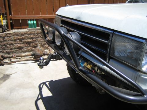 1990 Ford Bronco - front corner of a white SUV with a black tubular bumper and round lights, with an axle and gas bottles against a stone wall behind.