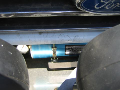 1990 Ford Bronco - Looking between two tires at a blue Accusump-style cylinder with a gauge mounted under a Ford tailgate bearing a chrome Ford emblem.