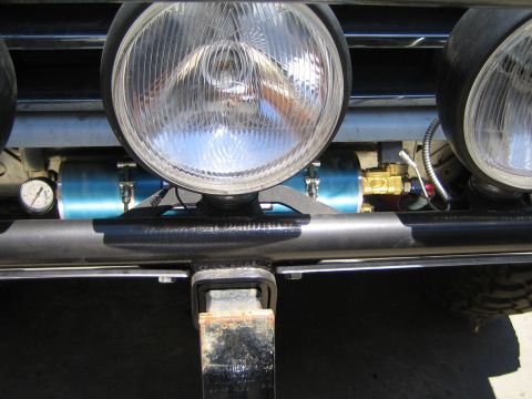 1990 Ford Bronco - close-up of round chrome driving lights mounted on a black tubular bumper, with a blue cylinder and a hitch receiver visible below.