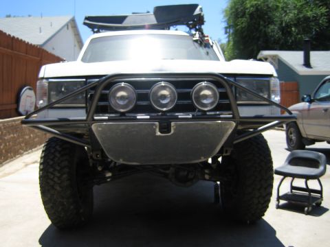 1990 Ford Bronco - head-on view of a white lifted SUV with a black tubular prerunner bumper, three round driving lights, and large mud tires.