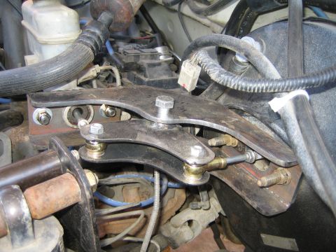 1990 Ford Bronco - Close detail of a custom dual stainless-steel bracket bolted across mounting studs in an engine bay, with blue plug wires and hoses behind…