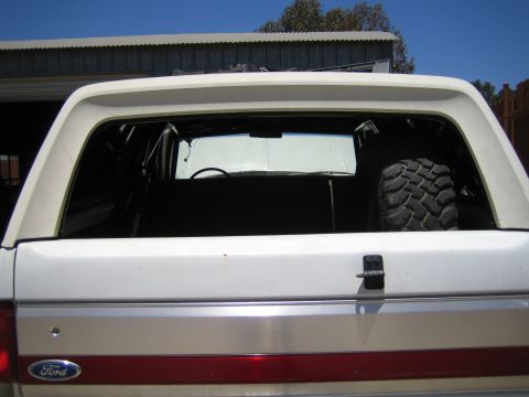 1990 Ford Bronco - Rear view of a white full-size SUV with a Ford oval, red taillight band and open rear glass revealing a mud tire inside, parked by a shed.