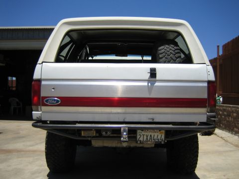 1990 Ford Bronco - Straight-on rear view of a lifted white Ford SUV with a Ford oval, red trim stripe, spare tire inside the cargo area and a tube rear bumper.