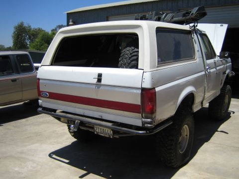 1990 Ford Bronco - Rear three-quarter view of a lifted white Ford full-size SUV with red trim stripe and large mud tires, parked beside another vehicle on a d…
