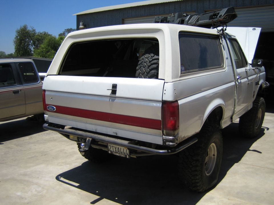 Three-quarter rear view of a lifted white Ford SUV on mud tires parked on a driveway by a metal shed