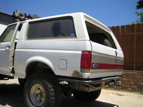 1990 Ford Bronco - Side rear quarter of a lifted white Ford SUV showing the red body stripe, Ford oval, fuel door, white wheel and chunky mud tire by a wood f…