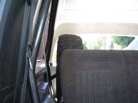 1990 Ford Bronco - Interior rear corner of an SUV showing a black roll bar, cloth bench seat back and a spare tire visible through the rear glass.