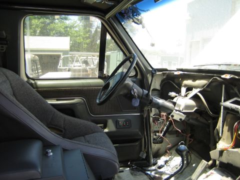 1990 Ford Bronco - Interior of a truck mid-disassembly with the steering wheel and column exposed, dash panels removed showing tangled wiring, and a cloth buc…