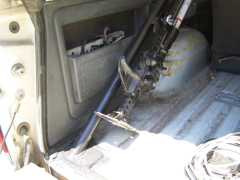 1990 Ford Bronco - Rear cargo area of a stripped SUV showing a black jack stored upright against a yellowed plastic fuel tank, bare metal floor and a coiled c…