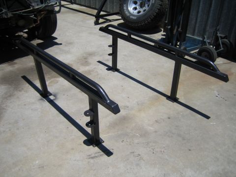 1990 Ford Bronco - pair of black tubular steel fabricated frame or rack rails resting on a concrete shop floor, with a tire and chassis parts behind them.