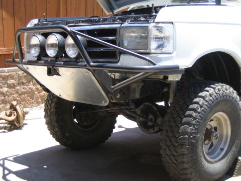 1990 Ford Bronco - Front of a white lifted Ford truck fitted with a black tubular prerunner bumper, three round driving lights and large mud tires, hood raise…