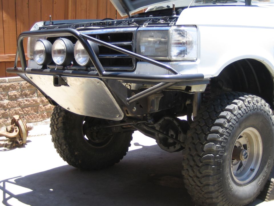Front of a white lifted Ford truck with a black tube bumper and three round driving lights, hood up