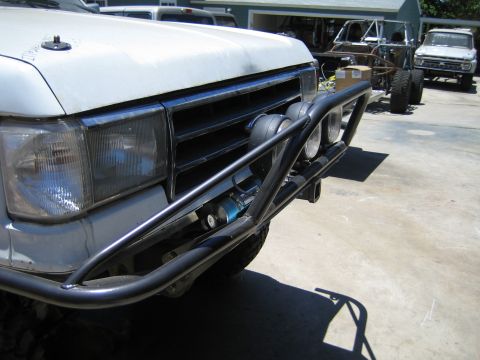 1990 Ford Bronco - Front corner of a white Ford truck showing the headlight, grille, black tubular bumper, driving lights and a blue coilover shock visible be…